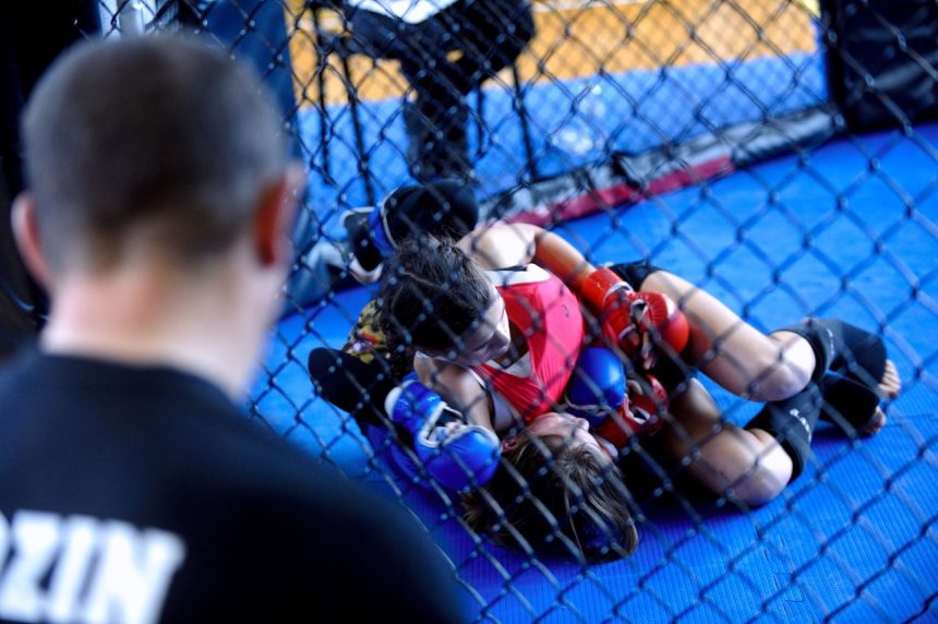 Duas lutadoras treinam MMA dentro de uma jaula de academia, enquanto um treinador observa.