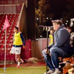 Treinador de futebol observa atentamente uma partida à noite, à beira do campo.