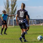 Jogadoras disputam uma partida de futebol feminino em um campo ao ar livre, sob sol.