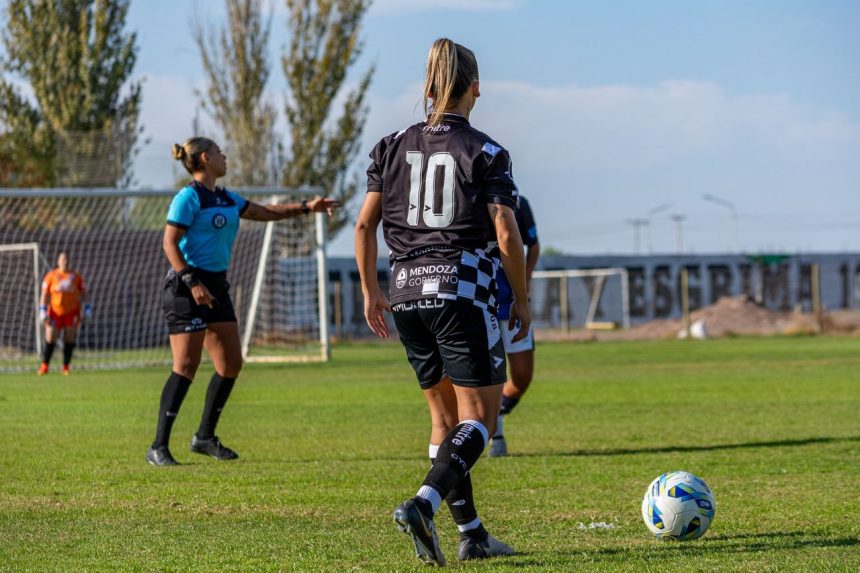 Jogadoras disputam uma partida de futebol feminino em um campo ao ar livre, sob sol.