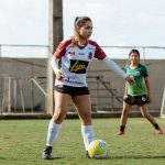 Duas adolescentes jogam futebol em um campo ao ar livre, usando roupas esportivas.