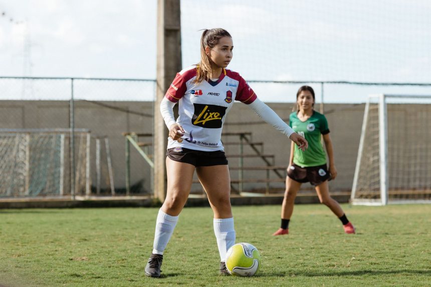 Duas adolescentes jogam futebol em um campo ao ar livre, usando roupas esportivas.